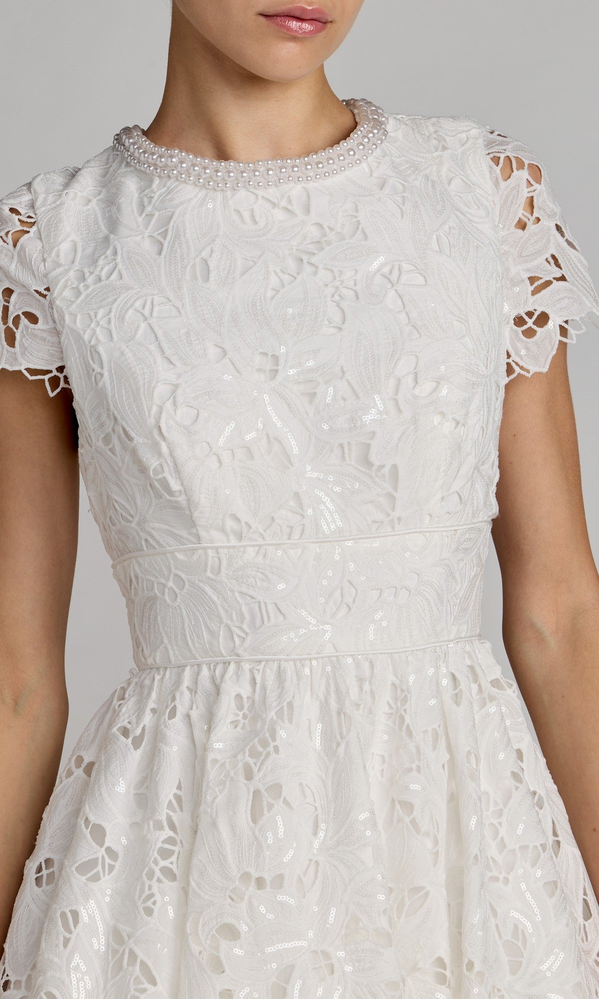 A woman wearing the Mac Duggal 56324 midi party dress is shown from the waist up. The white lace dress features short sleeves, a floral pattern, pearl trim at the neckline, and a fit-and-flare silhouette that flatters her figure.